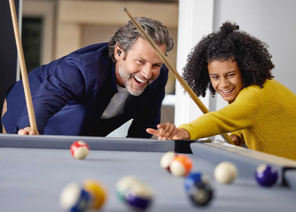 A man wearing Jabra Enhance™ Plus earbuds playing pool with a woman.